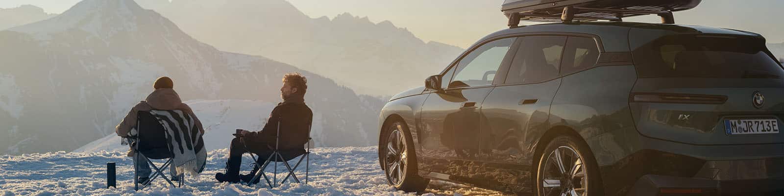 Zwei Personen sitzen auf Campingstühlen im Schnee neben einem BMW iX mit Dachbox und genießen den Blick auf verschneite Berge bei Sonnenaufgang.