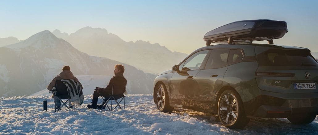 Zwei Personen sitzen auf Campingstühlen im Schnee neben einem BMW iX mit Dachbox und genießen den Blick auf verschneite Berge bei Sonnenaufgang.