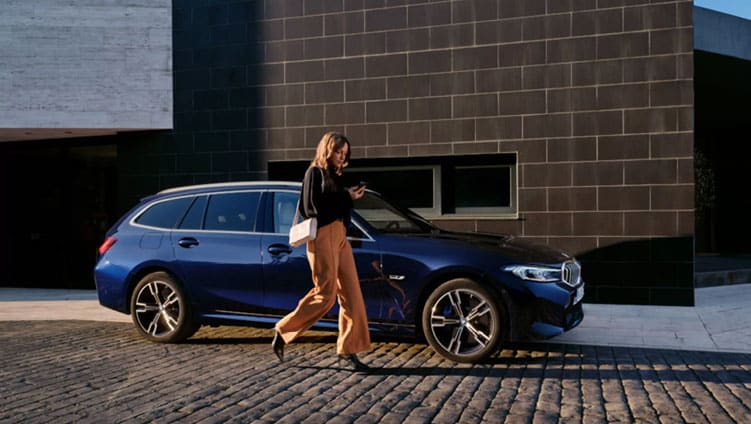 Frau geht neben einem dunkelblauen BMW Touring auf einem gepflasterten Weg.